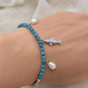 Könnte beinhalten: Ein Perlenarmband aus kleinen, runden, durchscheinenden blauen Steinen, die mit silberfarbenen Metallelementen durchsetzt sind. Das Armband ist mit zwei weißen, unregelmäßig geformten Süßwasserperlen und einem silberfarbenen Fisch-Anhänger verziert.
