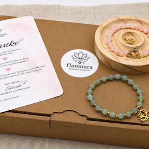 Puede incluir: Una caja de cartón marrón que contiene una tarjeta blanca con el texto Danke, una bandeja redonda de madera con una pulsera de cuentas rosas con dije de loto dorado, y una pulsera de cuentas verdes con dije de loto dorado, y cuentas doradas.