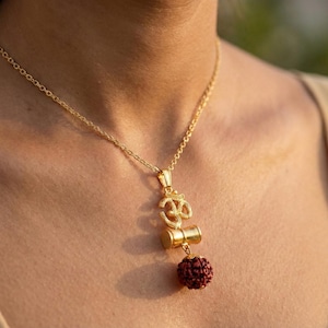 Puede incluir: Collar dorado con un colgante que presenta el símbolo Om, un elemento cilíndrico y una cuenta redonda de color marrón rojizo. La cadena es delicada y el colgante cuelga sobre un fondo neutro.