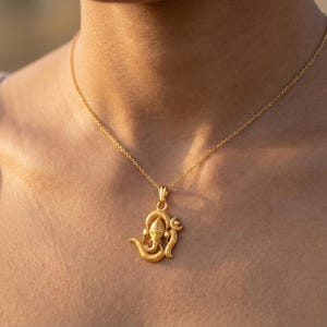 Może przedstawiać: Złoty naszyjnik z wisiorkiem z szczeg&oacute;łowym wzorem Ganesha. Wisiorek zawieszony jest na delikatnym łańcuszku. Naszyjnik jest noszony na tle odcienia sk&oacute;ry.