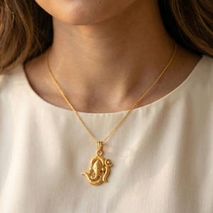 Może przedstawiać: Złoty naszyjnik z wisiorkiem przedstawiającym hinduskie b&oacute;stwo Ganesha. Wisiorek zawieszony jest na delikatnym łańcuszku. Naszyjnik jest noszony na kremowej bluzce. Biżuteria jest szczeg&oacute;łowa.