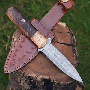 May include: A handcrafted knife with a patterned steel blade and a wooden handle. The handle is dark brown with white accents and a decorative emblem. The knife rests in a brown leather sheath, with a leather strap and a wooden base.