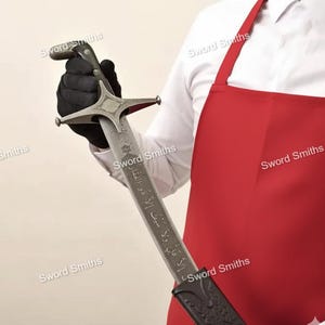 Può includere: Una spada d'argento con una guardia a forma di stella e un'impugnatura scura è tenuta da una persona con un guanto nero. La lama ha un testo inciso. La persona indossa una camicia bianca e un grembiule rosso. Le parole "Sword Smiths" sono stampate sull'immagine.