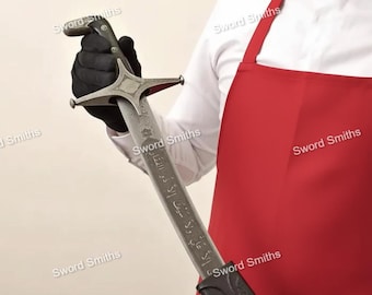 Pode incluir: Uma espada prateada com uma guarda em forma de estrela e um cabo escuro é segurada por uma pessoa com uma luva preta. A lâmina tem texto gravado. A pessoa está vestindo uma camisa branca e um avental vermelho. As palavras "Sword Smiths" são impressas na imagem.