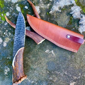 Puede incluir: Un cuchillo de caza artesanal con una hoja texturizada de color gris oscuro y un mango de asta natural. El cuchillo va acompañado de una funda de cuero marrón con costuras decorativas y una correa. El cuchillo y la funda descansan sobre una superficie gris texturizada.