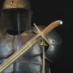 Puede incluir: Primer plano de una espada con empuñadura de cuero marrón claro y hoja plateada. La espada tiene una guarda en forma de cruz y se exhibe frente a una armadura. La hoja tiene un diseño grabado.