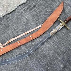 Puede incluir: Una espada curva con empuñadura de madera y guarda de latón, junto a su funda de cuero. La hoja tiene grabados decorativos. La funda es de cuero marrón claro con cinturón a juego.