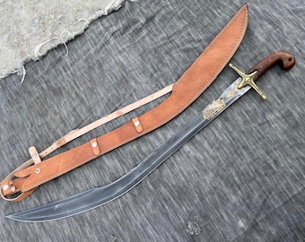 May include: A curved sword with a wooden handle and a brass crossguard, resting beside its leather sheath. The blade has decorative etchings. The sheath is tan leather with a matching belt.