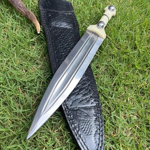 May include: A silver dagger with a bone handle and a black leather sheath rests on green grass. The dagger has a double-edged blade and a decorative handle. The sheath has a textured pattern.