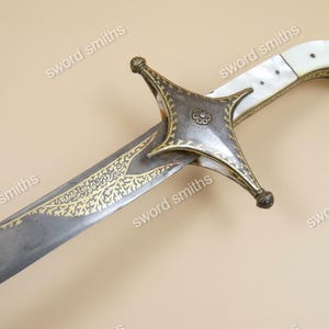 Op de afbeelding: Een antiek zwaard met een gebogen lemmet en sierlijke gouden details. De greep heeft een witte handgreep met een bloemmotief en een decoratieve pareerstang. Het lemmet heeft gouden versieringen.
