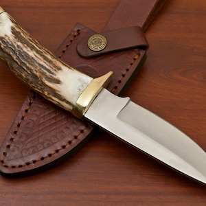 Könnte beinhalten: Ein Jagdmesser mit polierter Stahlklinge und einem Griff aus Geweih und Messing. Es liegt auf einer braunen Lederscheide mit geprägtem Muster und einem Messingknopf. Ein klassisches Design.