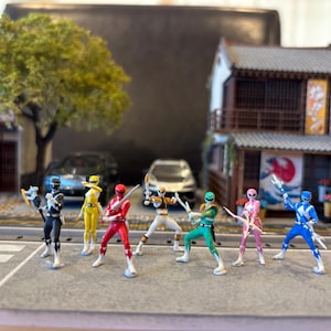 以下が含まれることがあります： 様々な色のパワーレンジャーのミニチュアフィギュアが、日本のスタイルの建物と車を背景にした通りのシーンにポーズをとっています。各フィギュアは武器を持っています。