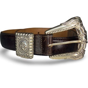 Op de afbeelding: Een bruine leren riem met een textuurpatroon en een zilverkleurige gesp. De gesp heeft een sierlijk ontwerp en de riem heeft een rechthoekige zilverkleurige lus met een kruisontwerp. De riem is ongeveer 3,8 cm breed.