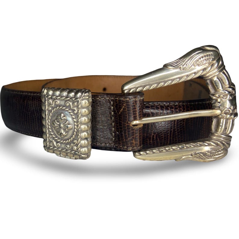 Op de afbeelding: Een bruine leren riem met een textuurpatroon en een zilverkleurige gesp. De gesp heeft een sierlijk ontwerp en de riem heeft een rechthoekige zilverkleurige lus met een kruisontwerp. De riem is ongeveer 3,8 cm breed.