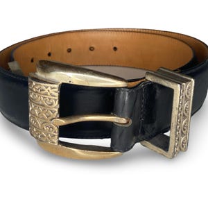 Op de afbeelding: Zwarte leren riem met een goudkleurige gesp met een sierlijk ontwerp. De riem heeft een lichtbruine binnenkant en een zilverkleurige rechthoekige gesp. Een klassiek accessoire.