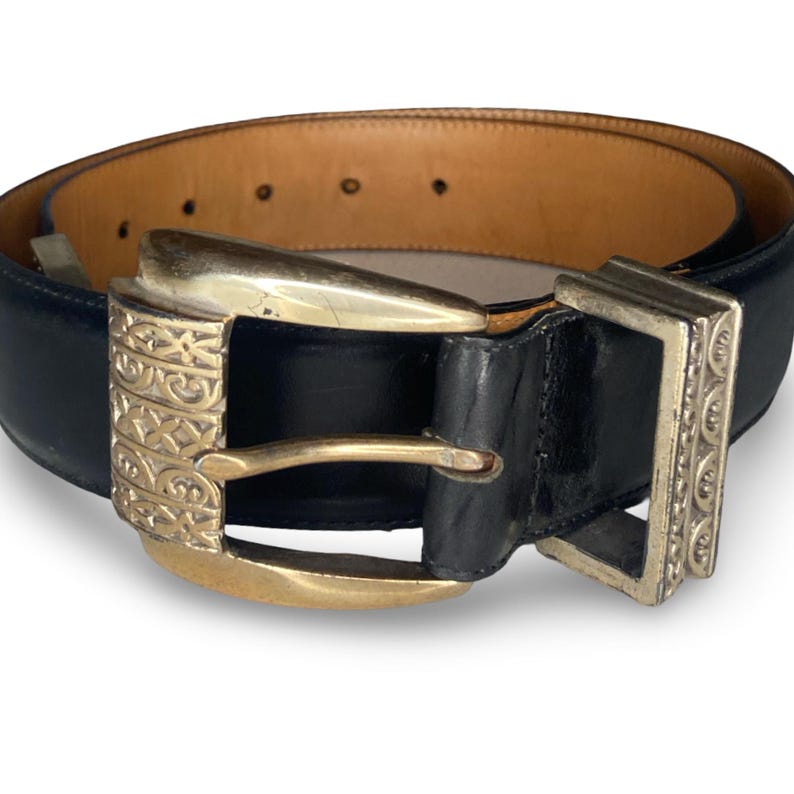 Op de afbeelding: Zwarte leren riem met een goudkleurige gesp met een sierlijk ontwerp. De riem heeft een lichtbruine binnenkant en een zilverkleurige rechthoekige gesp. Een klassiek accessoire.