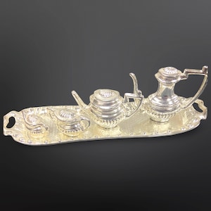 May include: A miniature silver tea set comprising a teapot, creamer, sugar bowl, and serving tray. The set features intricate detailing and a reflective surface. The tray is oval-shaped with handles. The set is likely a decorative item.