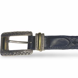 Op de afbeelding: Een marineblauwe leren riem met een zilverkleurige gesp en gouden accenten. De gesp heeft een decoratief, gestructureerd ontwerp. De riem is gevlochten en heeft een bijpassende zilveren en gouden riemlus.