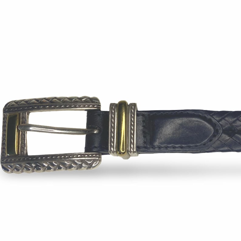 Op de afbeelding: Een marineblauwe leren riem met een zilverkleurige gesp en gouden accenten. De gesp heeft een decoratief, gestructureerd ontwerp. De riem is gevlochten en heeft een bijpassende zilveren en gouden riemlus.