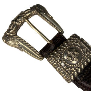 Op de afbeelding: Close-up van een bruine leren riem met een zilverkleurige gesp. De gesp heeft een sierlijk ontwerp met bloemen- en kruldetails. De riem is ongeveer 3,8 cm breed.