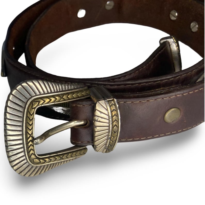 Op de afbeelding: Een bruine leren riem met een decoratieve zilveren en gouden gesp. De riem heeft een klassiek ontwerp met een metalen gesp en klinknagels. De riem is waarschijnlijk gemaakt van echt leer en is geschikt voor dagelijks gebruik.