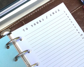 May include: A brown leather bound planner with a blue pen and a pink yarn ball. The planner has a white page with the text "50 BOOKS / 2022" and numbered lines from 1 to 25.