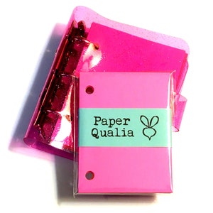 Op de afbeelding: Een roze notitieboekje met een turquoise label waarop "Paper Qualia" staat met een kleine konijnenillustratie. Het notitieboekje ligt op een roze glinsterende map.