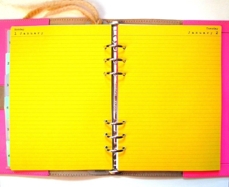 Op de afbeelding: Een gele, gestreepte planner met een zilveren binder. De linkerpagina is gelabeld "Maandag, 1 januari" en de rechterpagina is gelabeld "Dinsdag, 2 januari".