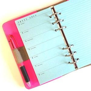 May include: A pink leather bound planner with a blue paper insert. The planner has a metal ring binding system and a pen holder. The paper insert has a weekly calendar layout with the month of April 2063 printed at the top.