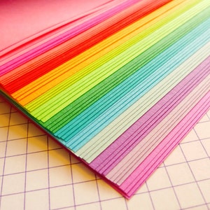 May include: A stack of colorful paper sheets in a rainbow pattern, arranged in a fan shape. The paper is folded and layered, creating a gradient effect from pink to purple.