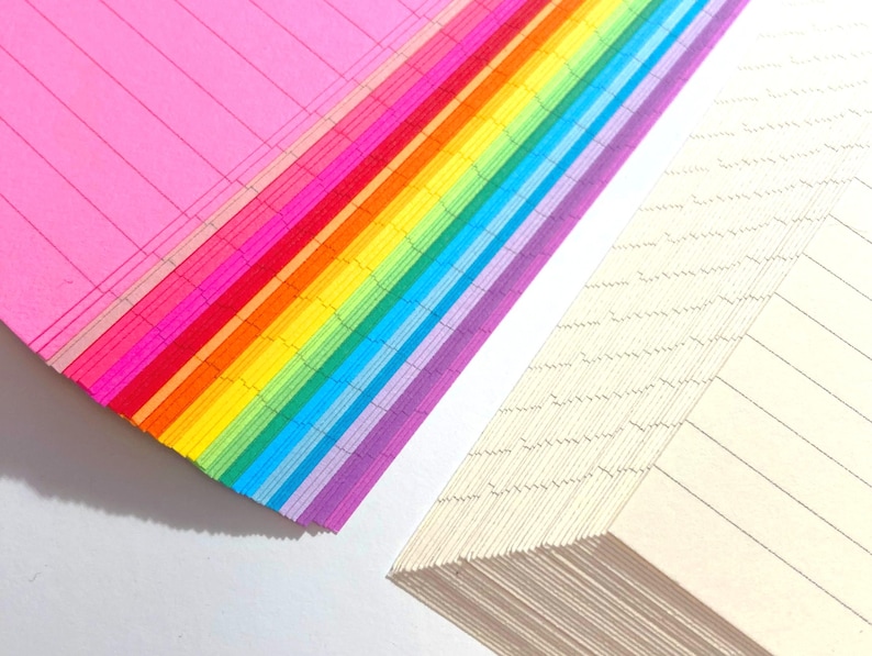 May include: A stack of colorful lined paper with a rainbow pattern. The paper is folded in half and the edges are visible. The paper is on a white surface.