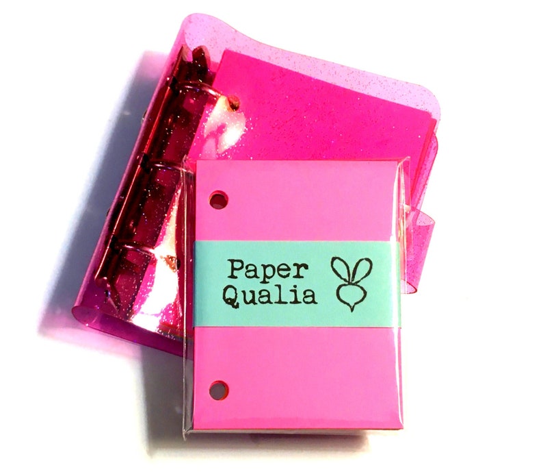 Op de afbeelding: Een roze notitieboekje met een turquoise label waarop "Paper Qualia" staat met een kleine konijnenillustratie. Het notitieboekje ligt op een roze glinsterende map.