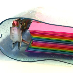Op de afbeelding: Een doorzichtige plastic map met een glinsterende afwerking en een regenboogstapel van kleurrijk papier erin.