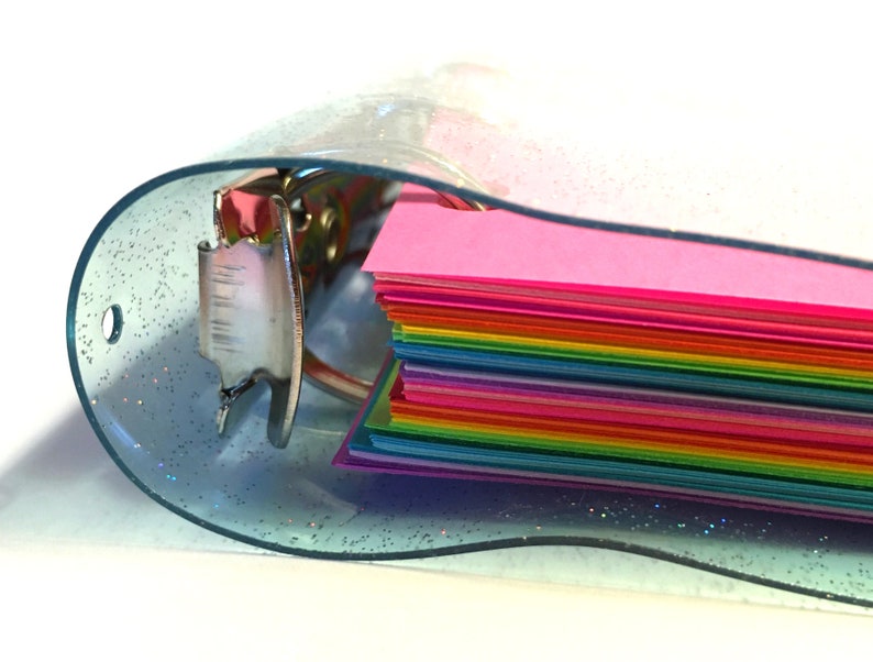 Op de afbeelding: Een doorzichtige plastic map met een glinsterende afwerking en een regenboogstapel van kleurrijk papier erin.