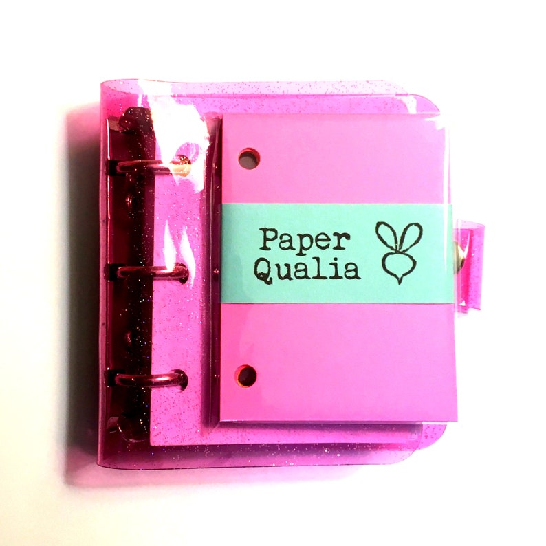 Op de afbeelding: Een roze glitter-omslagen map met een transparante plastic hoes. De map heeft een roze inlegvel met de tekst "Paper Qualia" en een klein konijnenlogo in een tealblauwe doos.