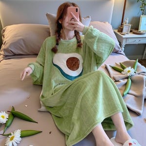 May include: A light green sleep dress with a large avocado graphic. The avocado has a brown pit and a white and blue outline. The dress is made of a soft, textured fabric and has a round neckline. The dress is a midi length.