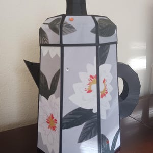 May include: A black and white floral patterned teapot with a black handle and spout. The teapot is made of paper and has a 3D design.