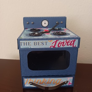 May include: A blue paper craft box shaped like a kitchen stove with a black oven window, a silver handle, and a stovetop featuring four burners and a clock. The front displays the text THE BEST Loved and Thinking.