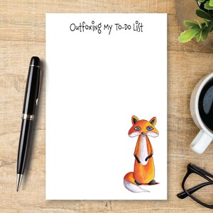 May include: A white rectangular notepad featuring the black printed text Outfoxing My To-Do List at the top and a colorful illustration of a standing orange fox with blue eyes and a white-tipped tail positioned in the bottom right corner.