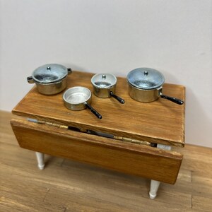 May include: A miniature wooden drop-leaf table with a natural finish, featuring four small metal cooking pots and pans on its surface. The set includes two lidded pots and two open pans, all with silver-toned bodies and black handles.