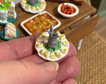 Puede incluir: Una figura miniatura de conejo gris centrada en una bandeja circular blanca rodeada de pequeños huevos rellenos cortados por la mitad con yemas amarillas, sobre una base de material verde texturizado, con el texto Renee Edmonston.