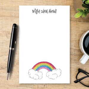 May include: A rectangular white notepad featuring the black text "Bright Ideas Ahead!" at the top and a colorful hand-drawn rainbow arching between two fluffy white clouds at the bottom, positioned next to a black pen and black glasses.