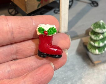 May include: A miniature red Christmas boot with a white fur trim and holly accents. The boot has a black sole and is a festive holiday decoration. The boot is a small, detailed ornament.