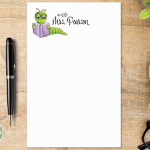 May include: A white rectangular notepad featuring a cartoon green caterpillar wearing glasses and reading a purple book, with the printed text "a note Mrs. Pearson" in black script. The notepad is positioned on a wooden surface with a pen.