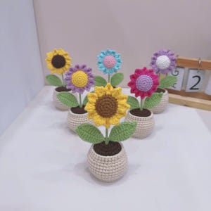 May include: A selection of handmade crochet sunflowers in small beige pots. The sunflowers feature yellow, purple, pink, and blue petals with brown centres. Each flower has green leaves and sits in a beige pot filled with brown soil.