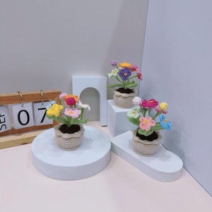 May include: Three crochet flower arrangements in small beige pots. Each pot contains colorful flowers in shades of pink, yellow, blue, and purple. The pots are displayed on white platforms, with a wooden calendar in the background.