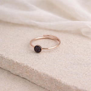 Könnte beinhalten: Ein roségoldener Ring mit einem dunkelgrauen, kreisförmigen Stein. Das Ringband umschließt den Finger, und der Stein ist mittig eingesetzt. Der Ring wird auf einer hellen, strukturierten Oberfläche präsentiert.