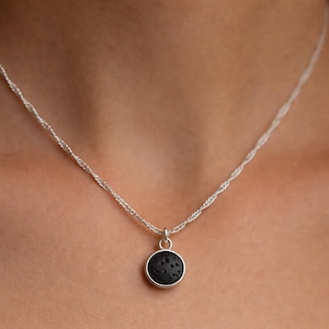 Puede incluir: Un collar de plata con un colgante redondo negro. El colgante tiene una superficie texturizada y está engastado en un bisel de plata. La cadena es de diseño trenzado y delicado. El collar se muestra sobre un fondo de tono de piel.