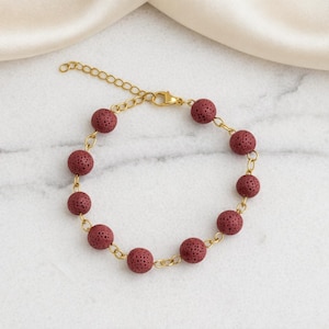 Può includere: Un braccialetto con catena dorata con perline di pietra lavica rosso scuro. Il braccialetto è esposto su una superficie di marmo bianco con un tessuto color crema sullo sfondo. Il braccialetto ha una catena regolabile. Le perline misurano circa 1 cm di diametro.