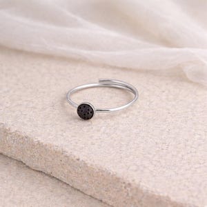 Può includere: Un anello argentato con una piccola pietra circolare scura. La sottile fascia dell'anello si avvolge attorno al dito. La pietra ha un aspetto strutturato e poroso. L'anello è mostrato su una superficie chiara e strutturata.